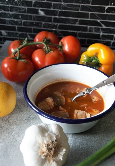 Foto weiß emailierte Schüssel mit blauem Rand und einer roten Fischsuppe daneben eine Knoblauchknolle Zitrone Tomate gelbe Paprika und einer Frühlingszwiebel